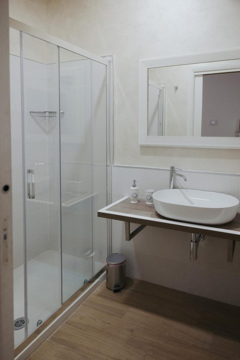 Bagno con box doccia in cristallo, lavabo ovale da appoggio su mensola in legno e specchio con cornice bianca