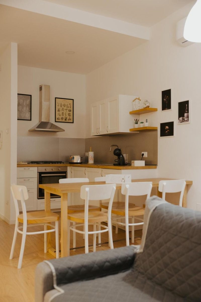 Il tavolo da pranzo con sei sedie bianche davanti alla cucina bianca a elle, con cappa in acciaio e mensole in legno