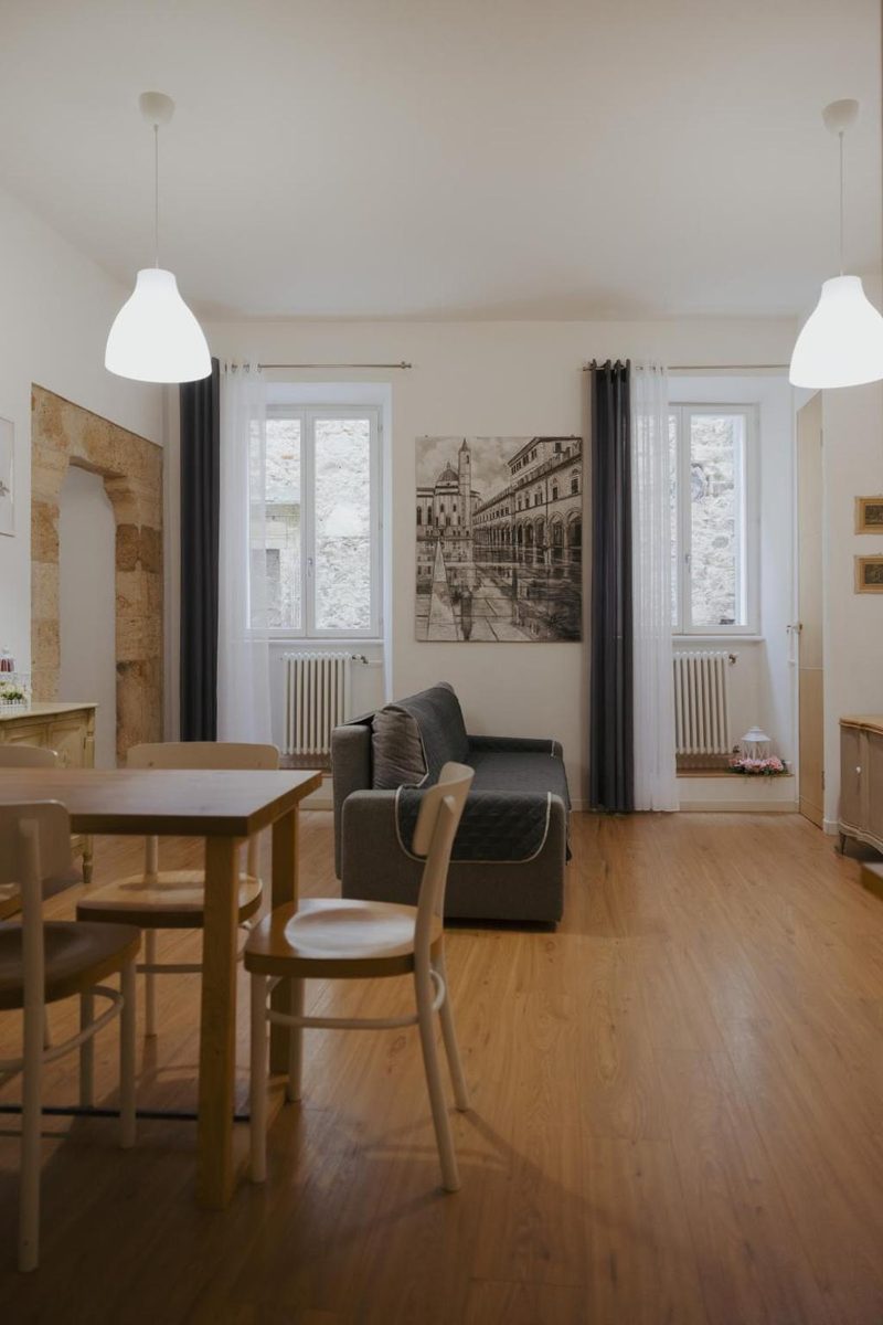 Zona giorno vista dal tavolo da pranzo: sedie in legno curvato, divano grigio, due finestre alte con radiatori in ghisa e la veduta di Ascoli stampata in bianco e nero alla parete